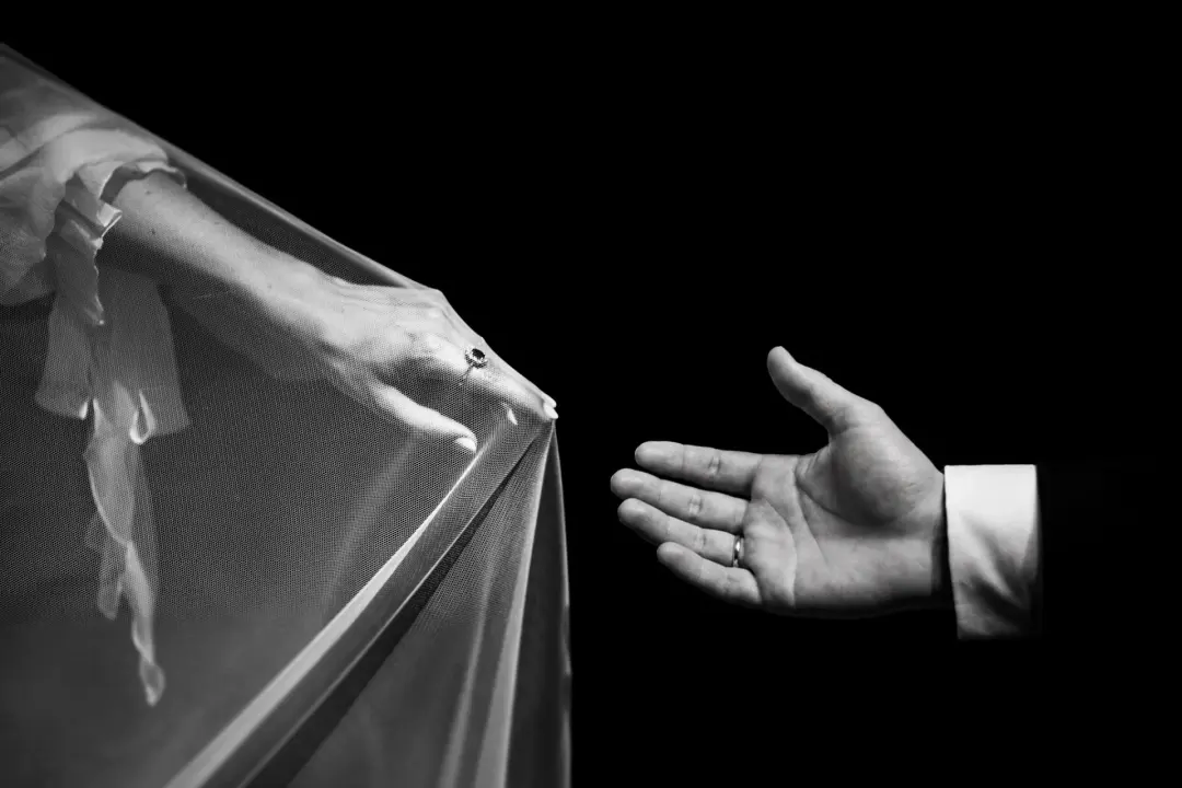Fotografía en blanco y negro de un detalle, con la mano del novio y el anillo de casado extendida hacia el borde del velo de la novia, sobre un fondo completamente negro que resalta las texturas del tul.