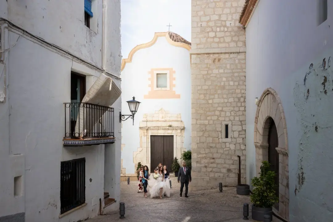La novia con su cortejo y su padre atraviesan una calle empedrada entre casas encaladas de un pueblo mediterráneo, con una iglesia de fachada barroca y un colchón asomando de un balcón al fondo, un detalle inesperado y curioso.