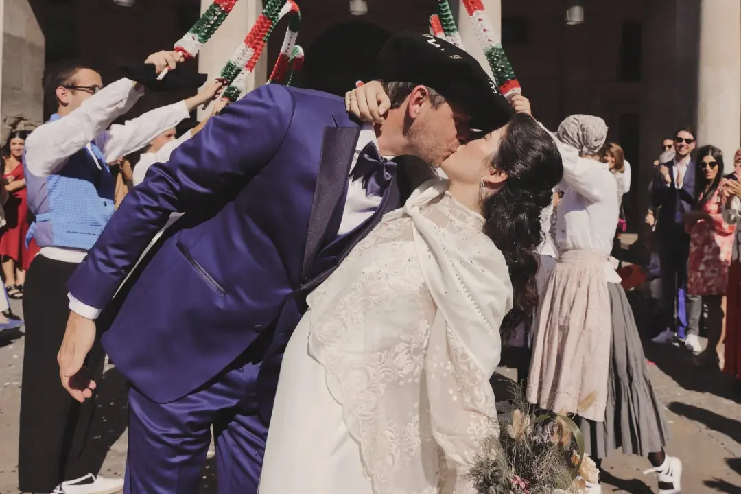 Los novios se besan a la salida de la iglesia mientras los invitados forman un arco con bastones festoneados que combinan el rojo, el blanco y el verde, y el novio lleva una boina tradicional bordada mientras ella sostiene un ramo silvestre de flores secas.